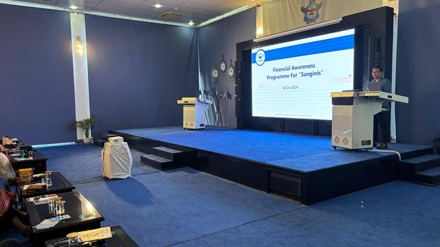 Auditorium stage with a title slide reading Financial Awareness Programme, with attendees seated to one side